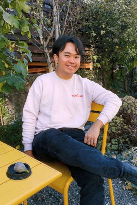 Patrick H. Pan smiling on a yellow chair in a garden, a fig on the table beside him